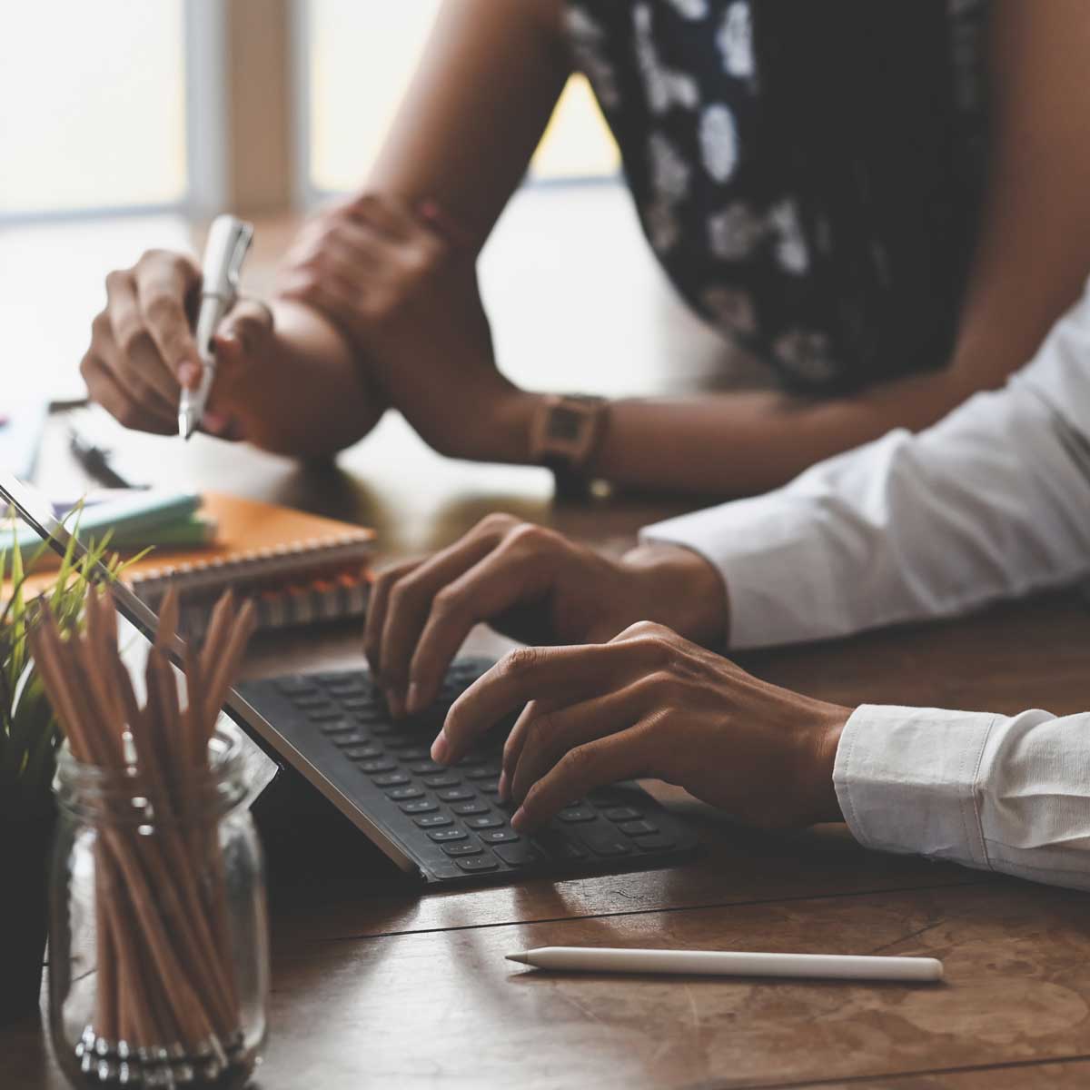 two employees working togethet on laptop and pen and pad. photo focuses on hands typing and writing with jar of pencils to left of center