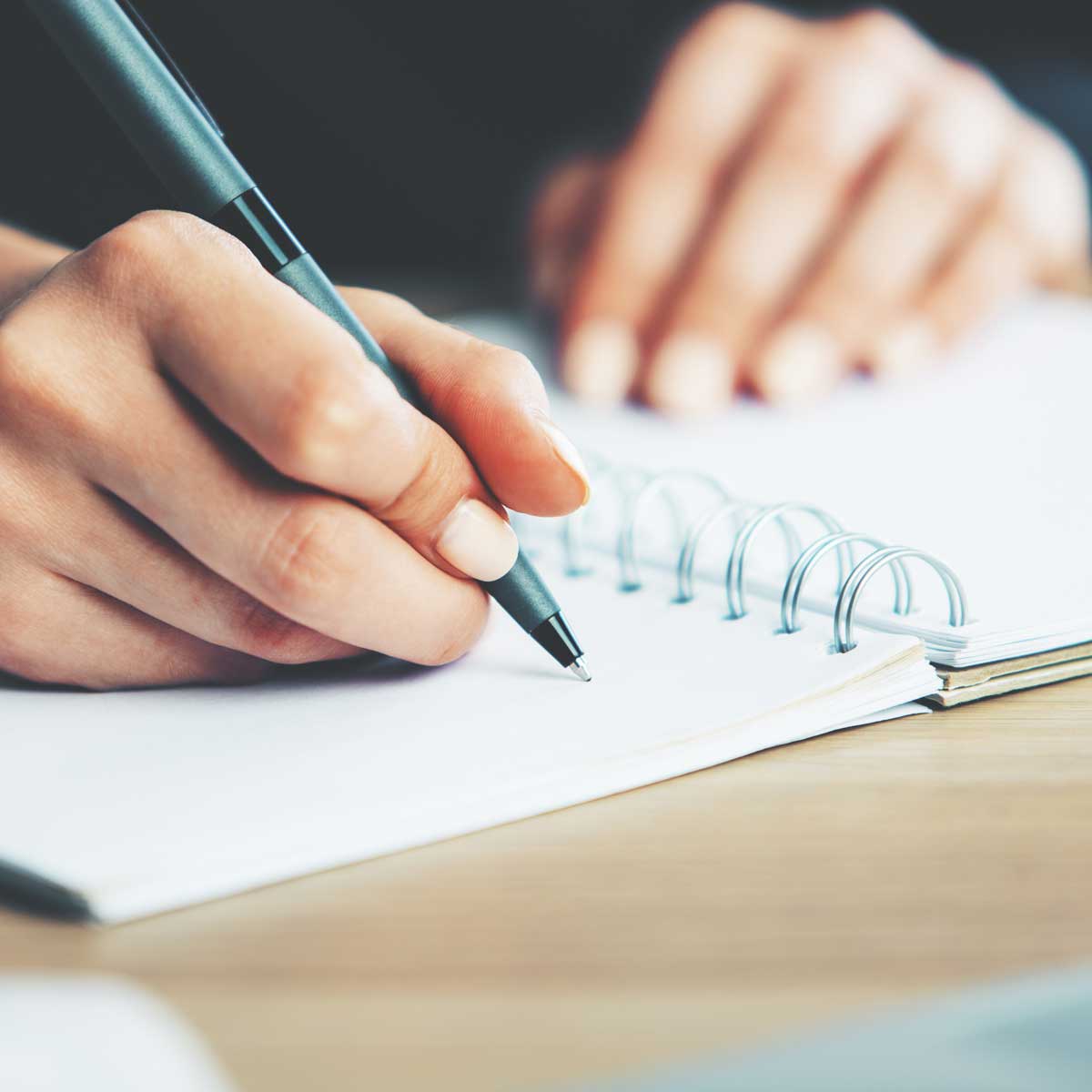 Woman writing in notepad; image focused on hand holding the pen on top of the spiral notepad