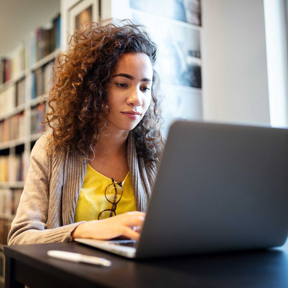 recent female college graduate sitting at a laptop analyzing student loan repayment options and plans and forbearance; sitting in a library with a book case behind her