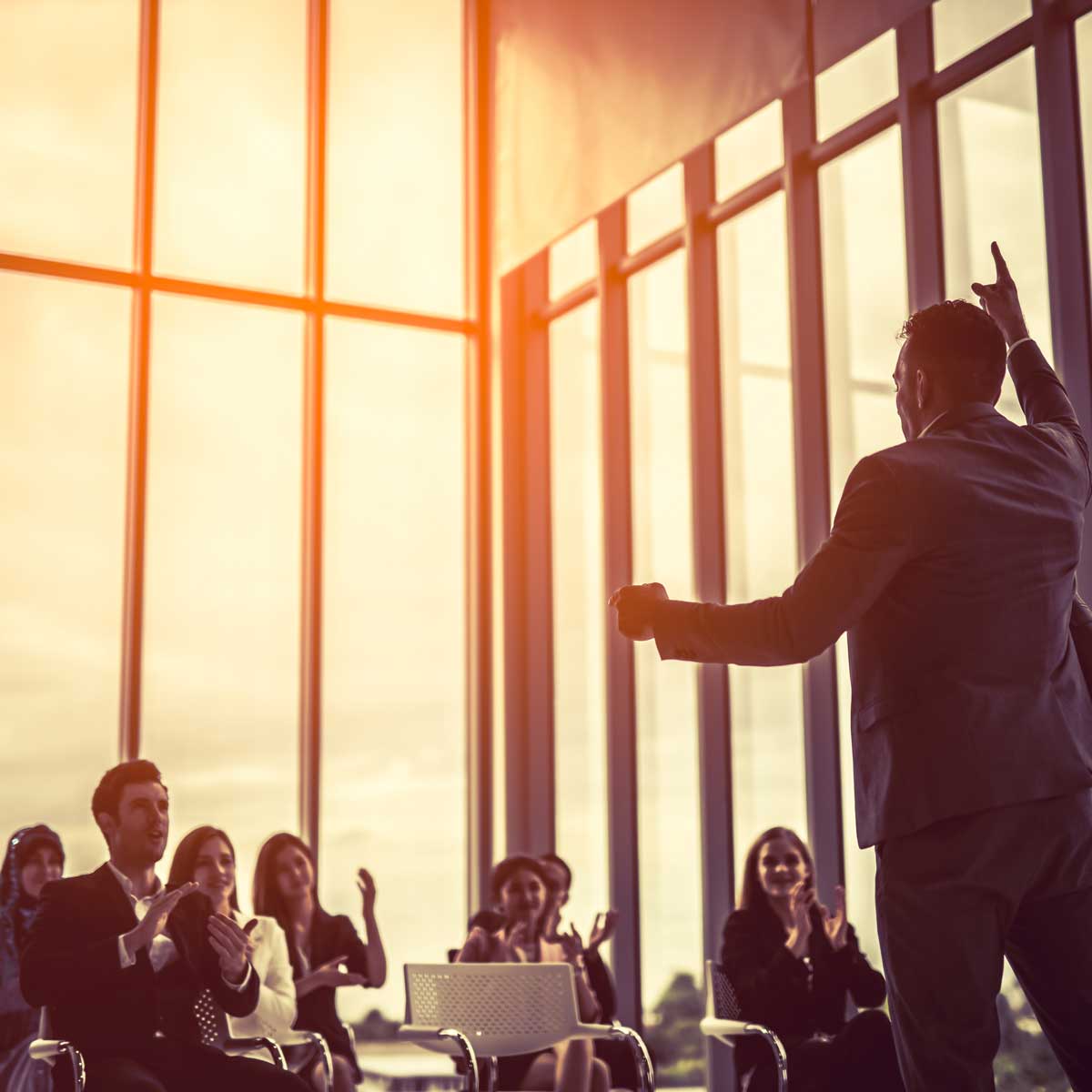 business leader presenting to crowd in room with glass windows and sun setting