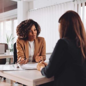 two female professionals in a job interview sitting across from each other at a table in a modern, open office